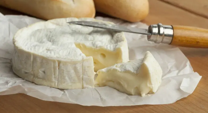 Un trozo cremoso de le camembert, con una rebanada cortada y lista para untar en pan crujiente.
