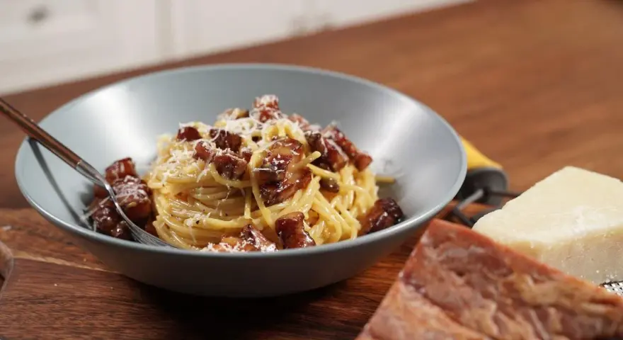 Miseczka spaghetti carbonara z boczkiem i parmezanem, obok kawałki sera i boczku.