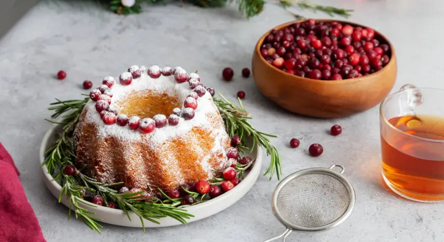 En vackert dekorerad sockerkaka med lingon och rosmarin, perfekt för julfika tips. En skål med lingon och en kopp te fullbordar julstämningen.