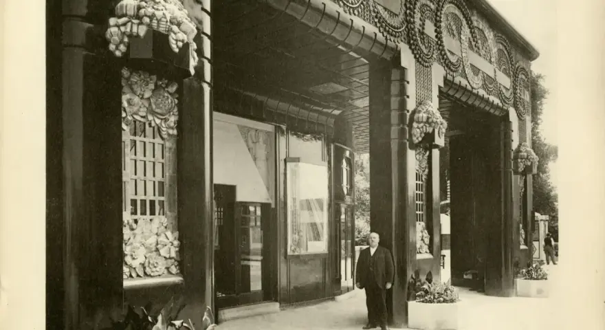 Façade de galerie de boutiques, style art déco, avec ornements floraux et motifs circulaires. Un homme en costume se tient sur le trottoir.