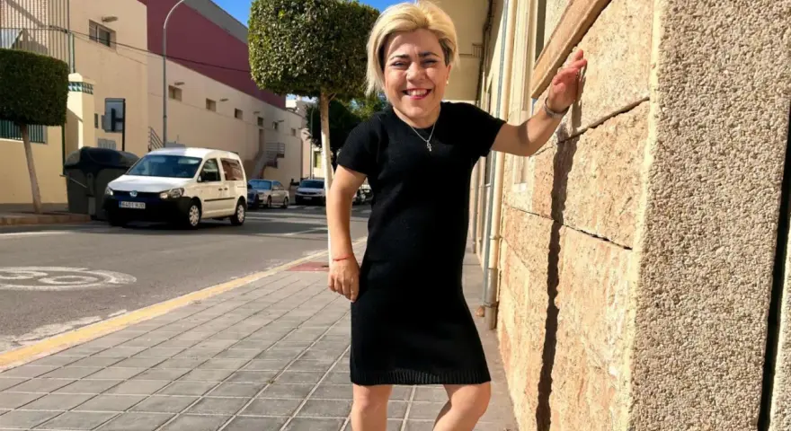 Mujer sonriente con vestido negro, apoyada en pared de piedra. Sus medidas cuerpo mujer son únicas y hermosas.