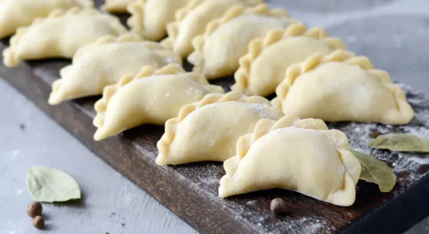 Surowe pierogi ułożone rzędami na drewnianej desce, posypane mąką.