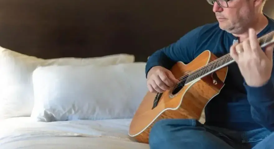 Mężczyzna w okularach gra na gitarze siedząc na łóżku. Gdzie ta keja nuty?
