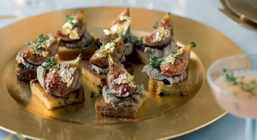 Deliciosos higos con foie sobre tostadas, adornados con pan de oro y tomillo, servidos en una bandeja dorada.