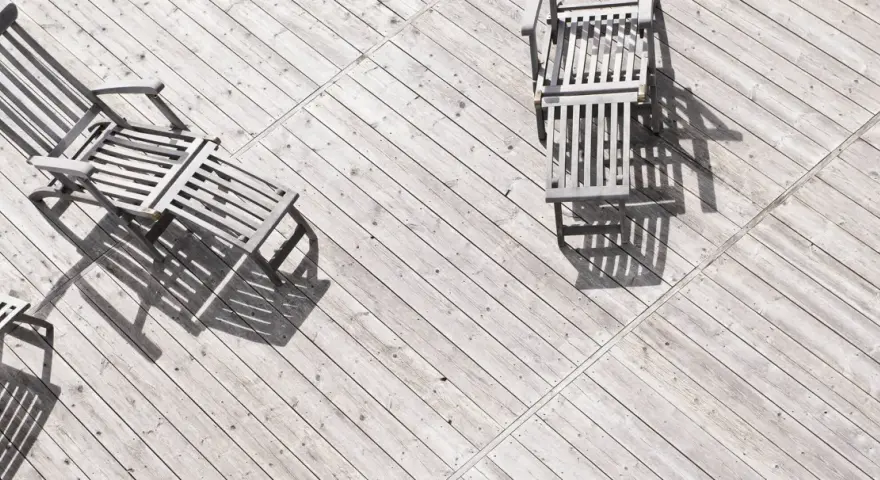 Tre solstolar i trä står på en sliten altan. Solen kastar långa skuggor över siox trall.