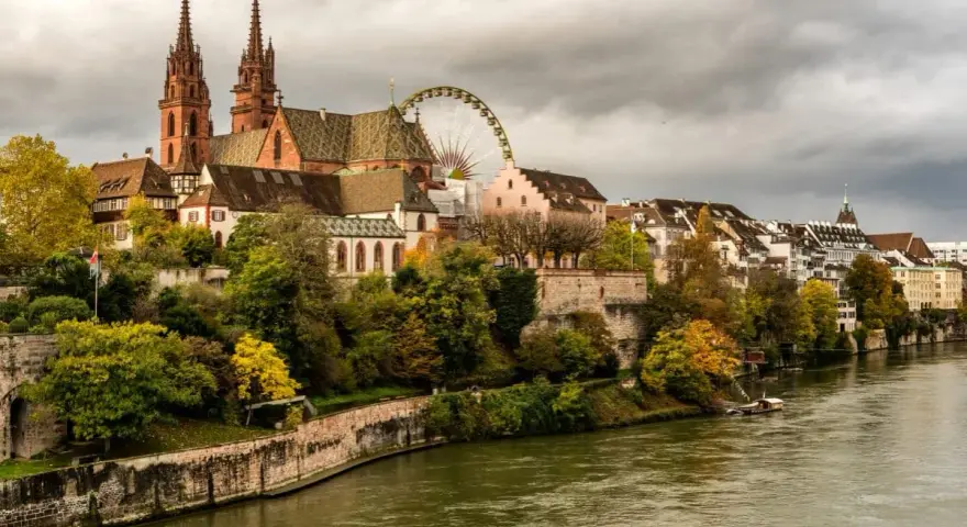 A scenic view of Basel's historic Minster and riverside buildings, perfect for a Basel itinerary. A Ferris wheel peeks out from behind the architecture.