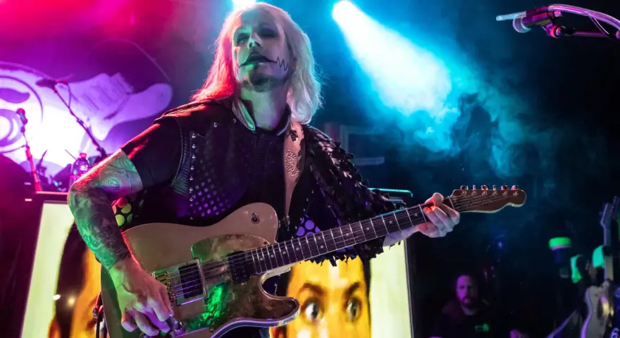John 5 mit seinem charakteristischen Make-up spielt Gitarre, im Hintergrund ein Bild von Marilyn Manson.