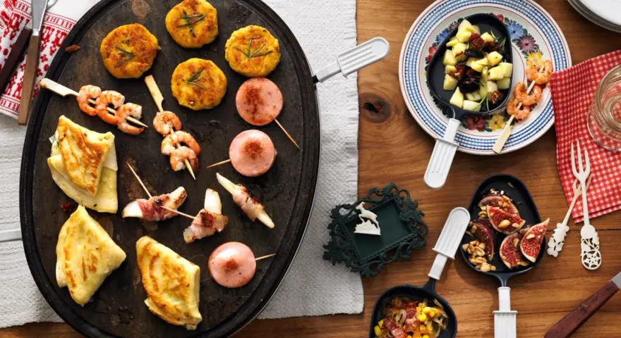 Raclette-Ideen für Kinder: Garnelen-Spieße, kleine Frikadellen, Käse-Speck-Röllchen und Crêpes auf einem Raclette-Grill.