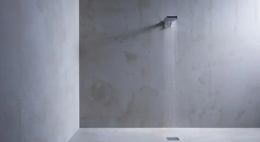 Douche minimaliste avec pommeau carré, eau tombant sur un sol en béton ciré sur galet. L'ambiance est épurée et moderne.