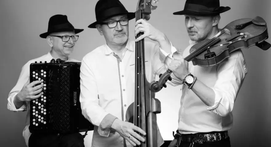 Trzech muzyków w czarnych kapeluszach grających muzykę klezmerską: akordeonista, basista i skrzypek.
