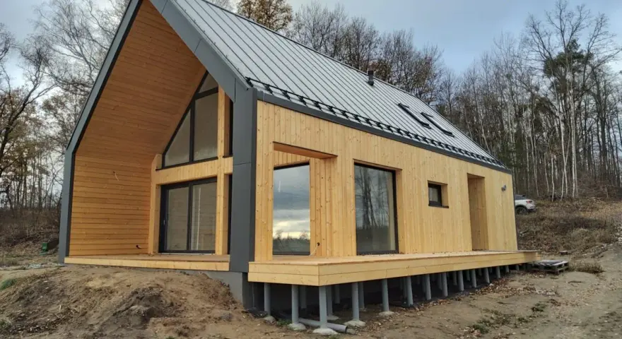 Nowoczesny dom 70m2 z drewnianą elewacją i dużymi oknami, otoczony lasem.