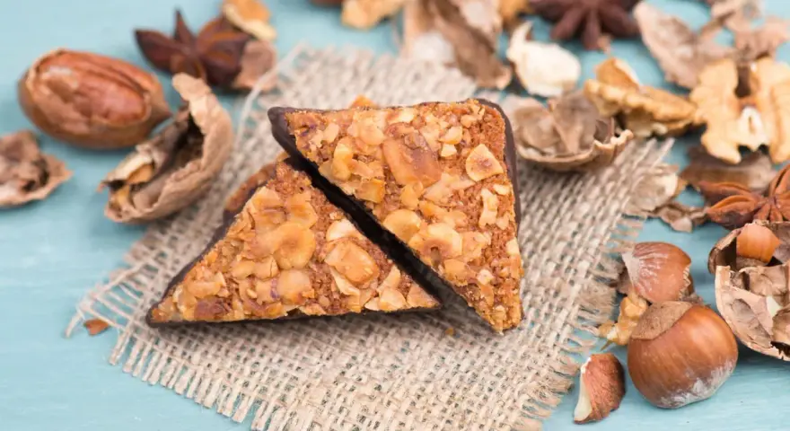 Zwei vegane Nussecken mit Schokoladenrand, bedeckt mit gehackten Nüssen, liegen auf einem Jutesack, umgeben von verschiedenen Nüssen und Sternanis.