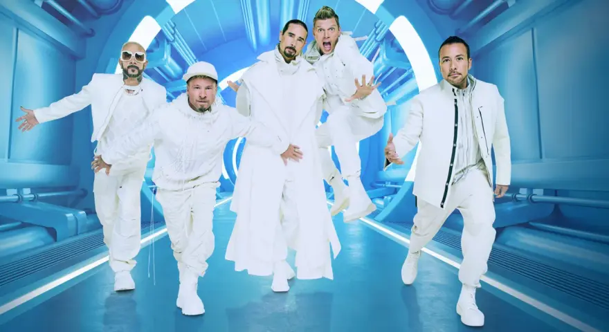 Backstreet Boys w białych strojach, gotowi na koncert. Ich energia jest zaraźliwa, a futurystyczna scenografia podkreśla ich styl.