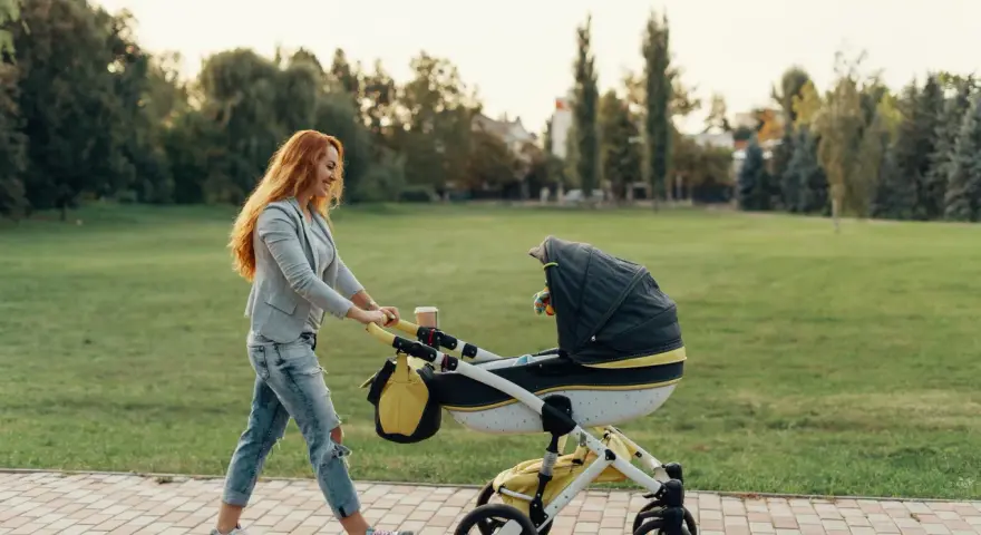 Ruda kobieta z uśmiechem pcha wózek podczas pierwszego spaceru z noworodkiem w parku.