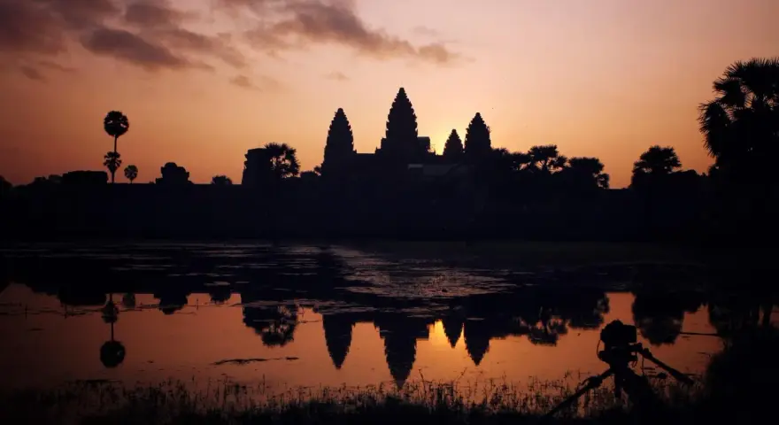 Zachód słońca nad Angkor Wat, idealne tło dla niezapomnianych kambodżańskich wakacji. Widok odbija się w wodzie, a na pierwszym planie statyw z aparatem.