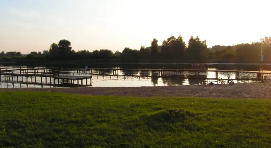Drewniane pomosty na spokojnym jeziorze starogrodzkim, z piaszczystą plażą i zieloną trawą w tle.