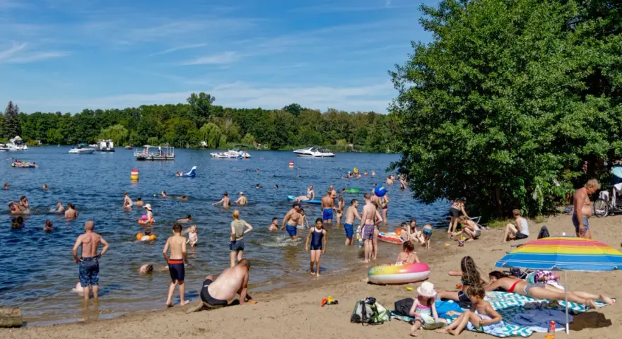 Menschen genießen das Reschensee baden an einem sonnigen Tag. Kinder planschen, Erwachsene schwimmen und entspannen am Ufer. Boote gleiten über das Wasser.