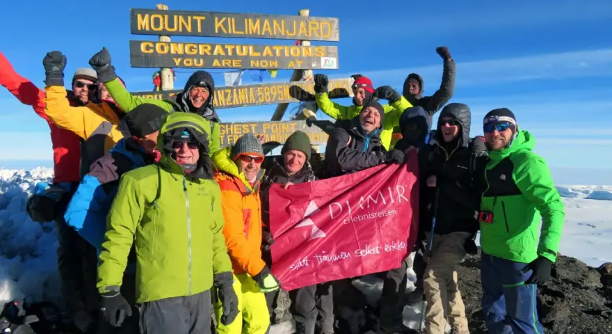 Glückliche Gruppe feiert auf dem Gipfel des Kilimandscharo. Sie haben den Gipfel erreicht und halten ein Banner hoch. Ein unvergessliches Erlebnis, den Kilimandscharo zu besteigen!