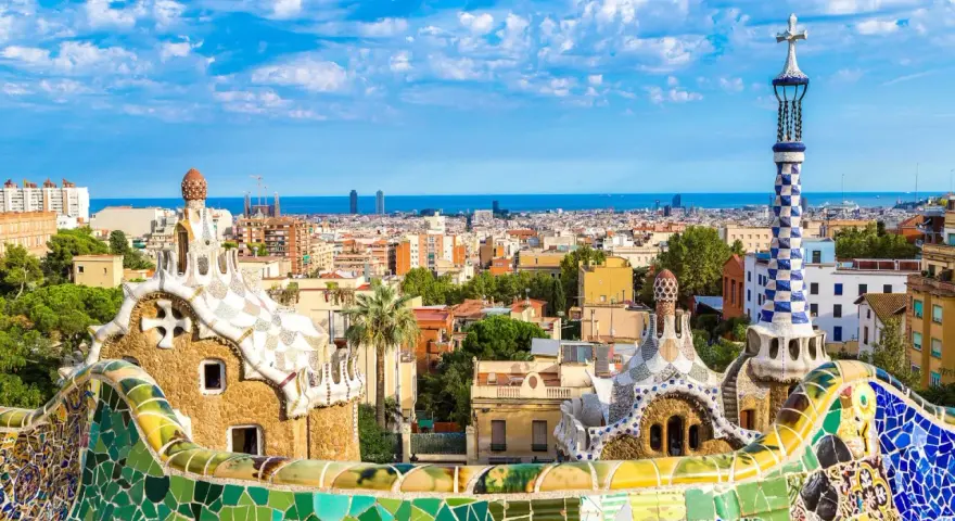 Kolorowe mozaiki i fantazyjne budowle Parku Güell, dzieło Antonio Gaudiego, na tle błękitnego nieba i Barcelony.