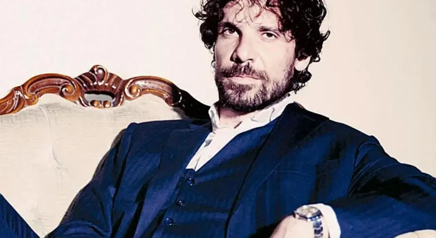L'attore Montanari, con barba e capelli ricci, indossa un completo blu e una camicia bianca, seduto su una poltrona d'epoca.