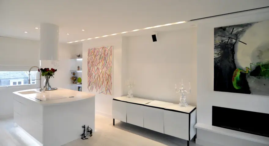 Modernt kök och vardagsrum med vita ytor, konst och spotlights som skapar en ljus och inbjudande atmosfär.