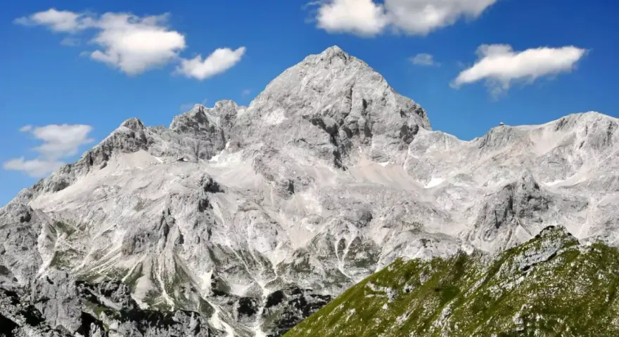 Majestätischer Triglav im Sonnenschein, bereit für die Triglav-Besteigung. Grüne Hänge und Felsen unter blauem Himmel mit Wolken.