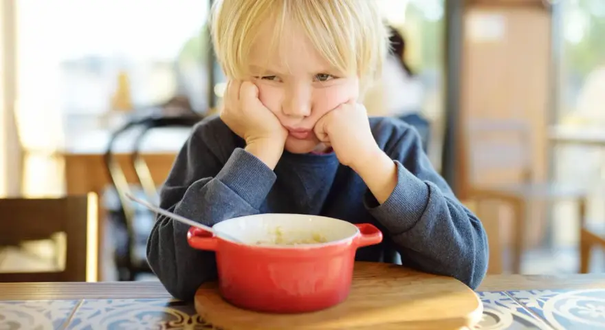 Ein Junge mit blonden Haaren stützt seinen Kopf auf die Hände und schaut lustlos auf eine Schüssel Porridge. Er isst nicht, obwohl er 10 Jahre alt ist.