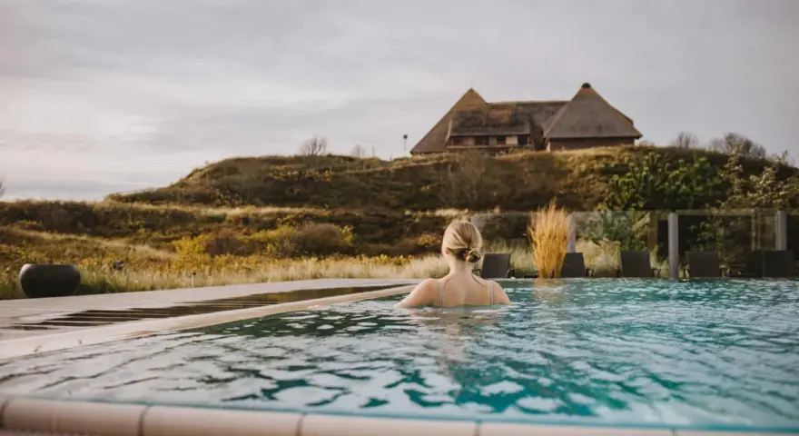 Frau genießt entspannten Wellness Tag in Schleswig-Holstein. Ein idyllischer Pool mit Blick auf Reetdachhaus und Dünenlandschaft.