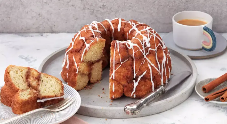 Ein köstlicher Monkey Bread mit Zimt und Zuckerguss, bereit zum Teilen. Daneben eine Tasse Kaffee und Zimtstangen.