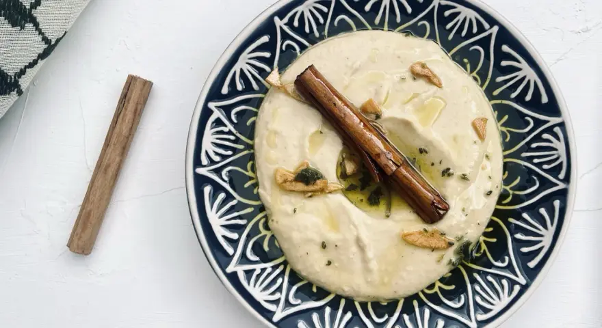 Cremiger Hummus, selbst gemacht, mit Zimtstange und Knoblauch garniert auf einem blau-weißen Teller.
