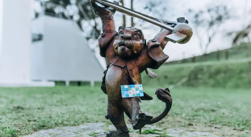 Figurka lew hewelion z kartą miejską w łapie, stojąca na bruku wśród zieleni.