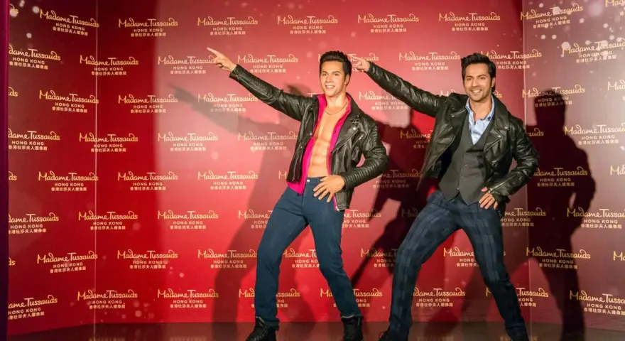Dwie woskowe figury, jedna z odkrytym torsem, stoją na tle powtarzającego się logo Madame Tussauds w Hongkongu.