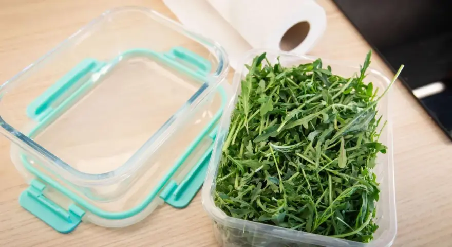 Frischer Rucola in einer Plastikschale neben einem leeren Glasbehälter. So gelingt das Rucola aufbewahren am besten.