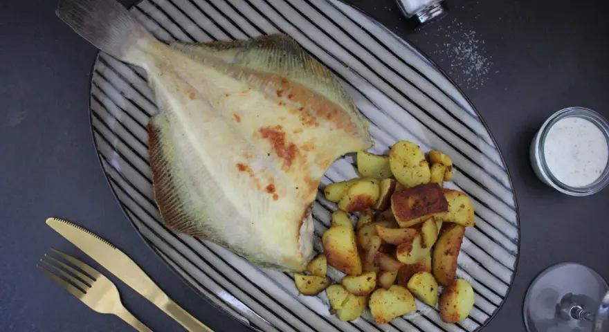 Gebratene Scholle Ostsee mit Bratkartoffeln und Dip auf einem gestreiften Teller, daneben ein Salzstreuer und goldenes Besteck.