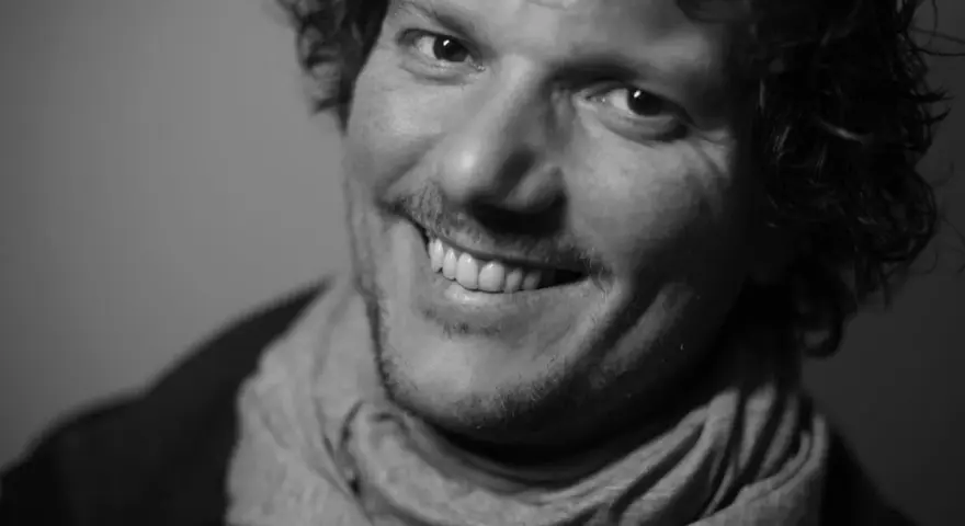 Uśmiechnięty Filip Ćwik w szarym szaliku, z kręconymi włosami i zarostem.