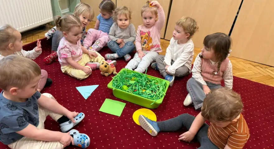 Dzieci siedzą na dywanie wokół zielonego pojemnika z zieloną trawą i jajkami.