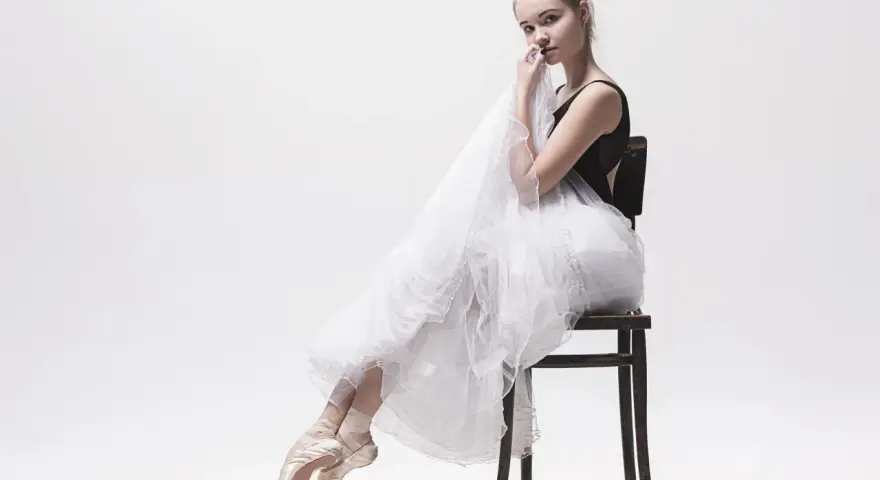 Młoda tancerka w baletkach i tiulowej spódnicy, uosobienie stylu balletcore, siedzi na krześle, trzymając w dłoniach delikatną tkaninę.
