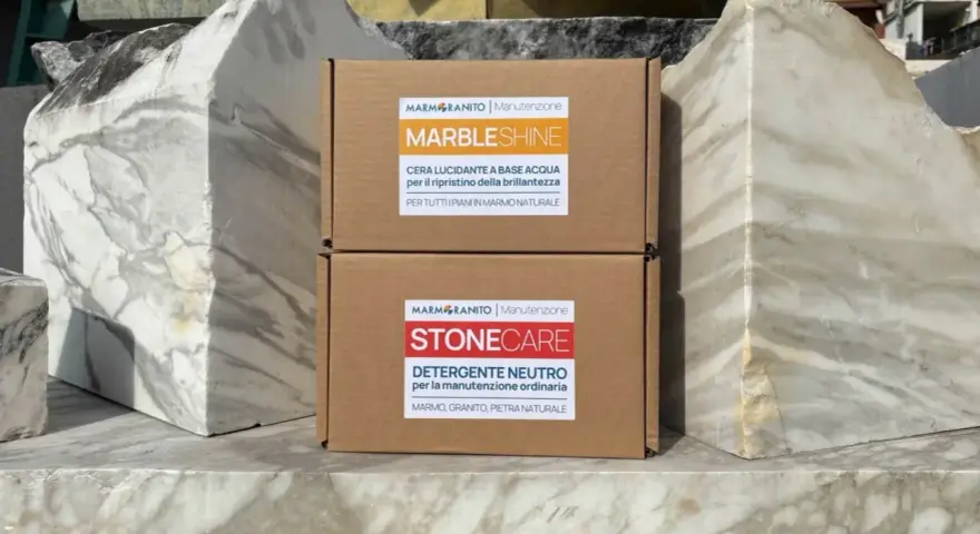 Due scatole di prodotti per la pulizia del marmo, "Marble Shine" e "Stone Care", appoggiate su blocchi di marmo grezzo.
