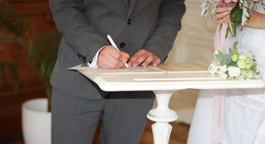 Pan młody podpisuje dokumenty, co się mówi na ślubie cywilnym? W tle panna młoda z bukietem.