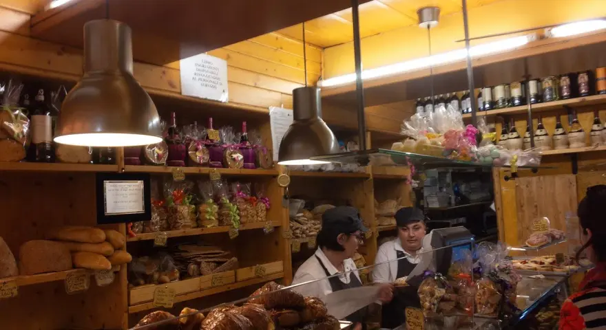 Interior of a bakery with shelves of bread, pastries, and bottles. Two women are behind the counter.