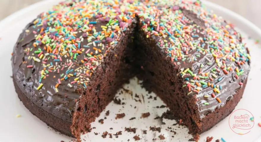 Ein saftiger, veganer Kuchen mit Schokoladenglasur und bunten Streuseln. Ein Stück fehlt, was Lust auf mehr macht.