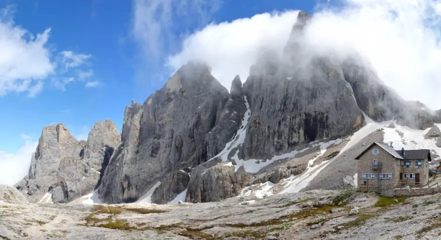 Rifugio in pietra ai piedi di imponenti vette rocciose, con neve residua e cielo azzurro. Un luogo incantevole al passo Valles.