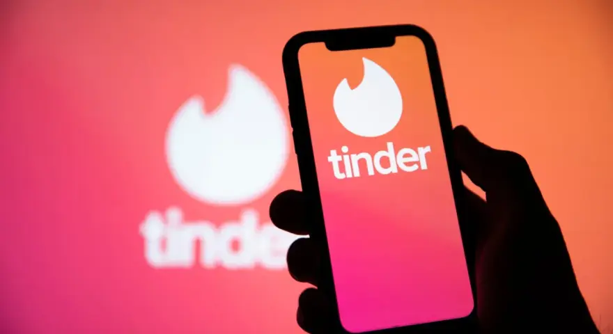 Dłoń trzyma smartfon z logo Tinder. W tle widać większe, rozmyte logo aplikacji, sugerujące nowe tinder powiadomienia.