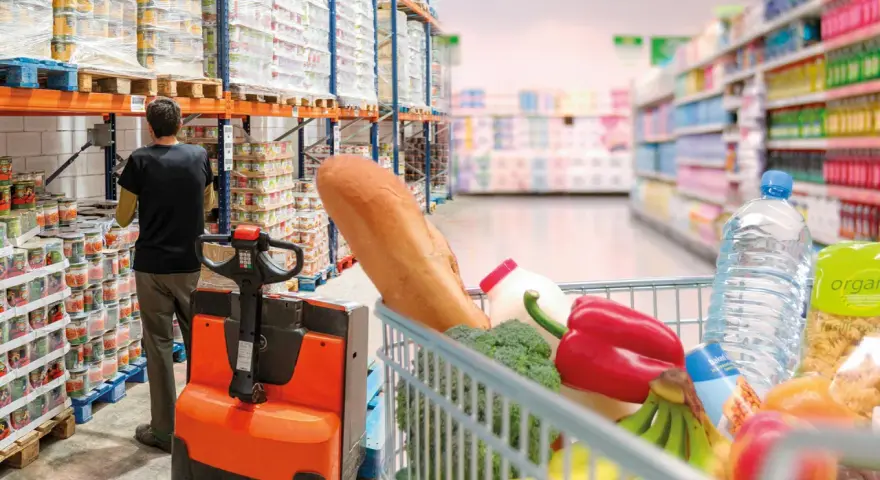 W magazynie FMCG, pracownik obsługuje wózek widłowy, podczas gdy wózek sklepowy pełen produktów czeka na zakupy.