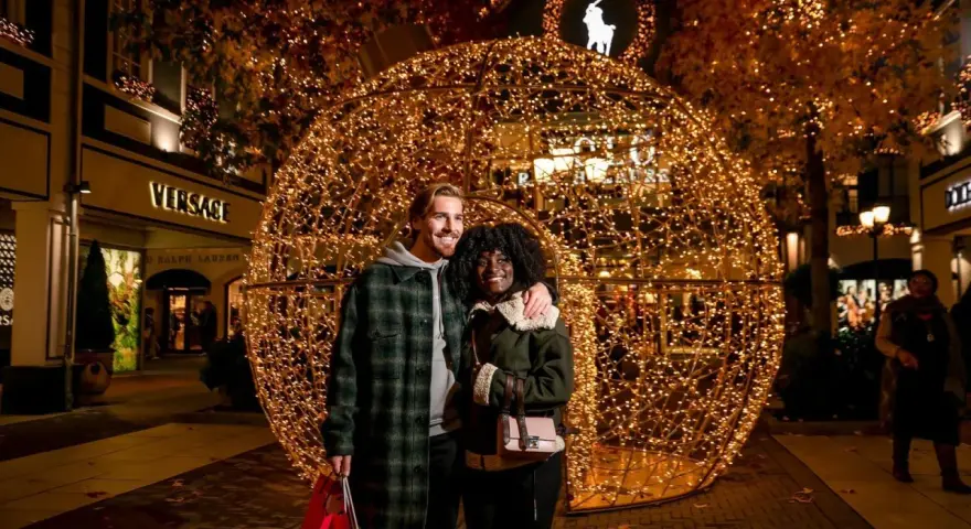Ein glückliches Paar vor einer riesigen Lichterkugel auf dem weihnachtsmarkt roermond. Geschäfte wie Versace und Ralph Lauren sind im Hintergrund zu sehen.
