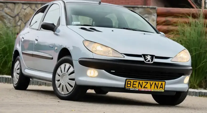 Jasny Peugeot 206 1.4 benzyna z żółtą tablicą "BENZYNA" na zderzaku, zaparkowany na zewnątrz.