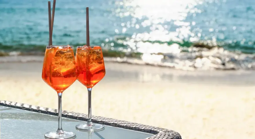 Zwei Aperol Spritz Gläser mit Eis und Orangenscheiben stehen am Strand. So trinkt man Aperol Spritz in Italien!