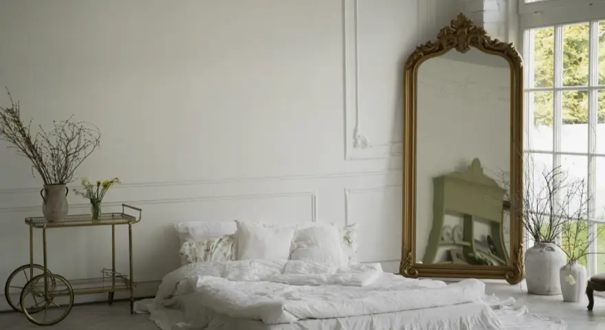 Elegancka sypialnia z łóżkiem w bieli, ozdobiona sztukaterią przypominającą lamperię angielską. Złote lustro i wózek dodają blasku.