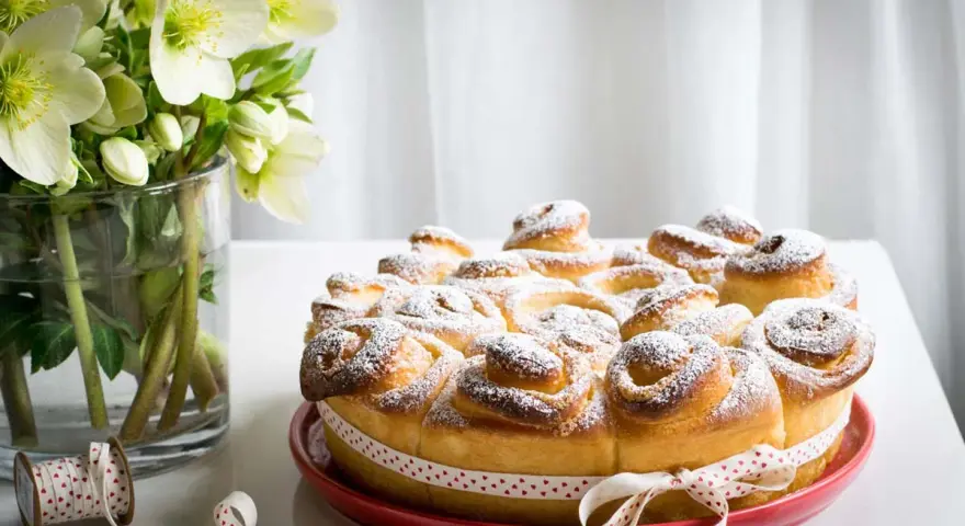 Torta di rose soffice e dorata, cosparsa di zucchero a velo, decorata con un nastro a cuoricini. Accanto, un vaso con fiori bianchi.