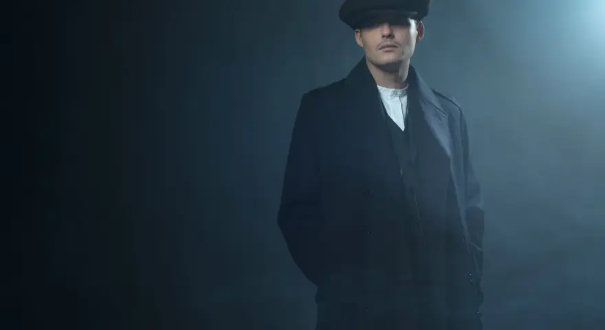 Mężczyzna w kaszkiecie i płaszczu, z charakterystycznym wąsem, emanuje chłodnym spojrzeniem w stylu Peaky Blinders.